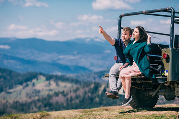 Side view of a smiling woman embracing man against  offroad old jeep and mountains. Woman in dress. Man show on landscape. Blue sky sunny day. Amazing travel