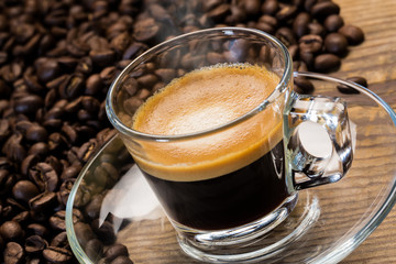 A transparent glass cup of hot steamy espresso on a saucer standing on a wooden table top, surrounded by coffee beans