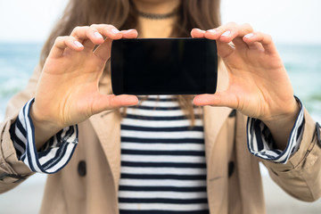 Woman in beige coat shows the smart phone screen on the beach