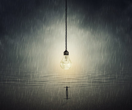 Surreal backround of a man standing with wide opened hands in front of a huge bulb near the ocean in a rainy day. Idea concept