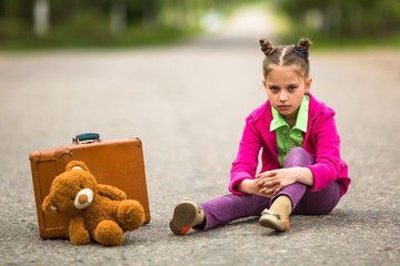 Little girl sitting on the road with a suitcase and a Teddy bear.