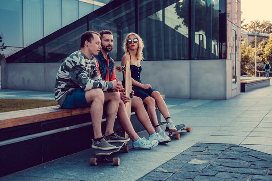 Two hipster males and blond female with longboards resting.