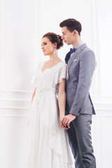 Gorgeous bride in white dress and handsome groom in grey tuxedo posing in retro interior.