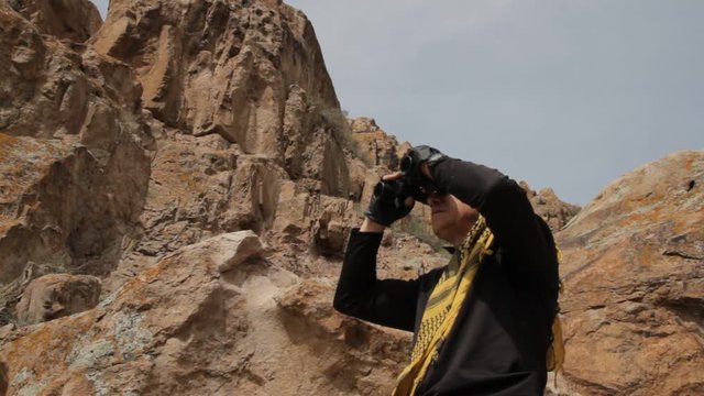 A man dressed in Bedouin shawl looks through binoculars among the rocks