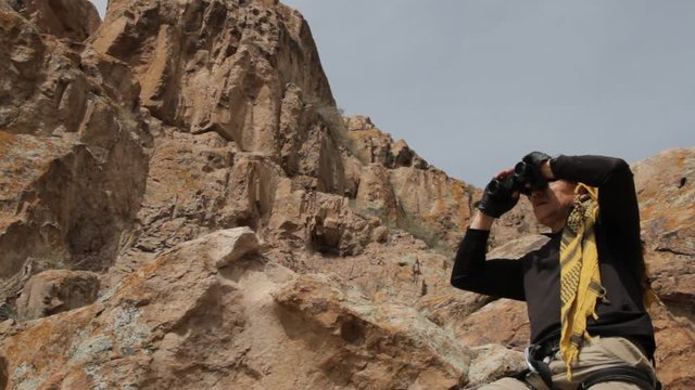 A man dressed in Bedouin scarf looking through binoculars among the rocks