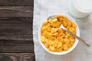 Tasty corn flakes in bowl with bottle of milk. Rustic wooden background with homespun napkin. Healthy crispy breakfast snack. Place for text. Top view, flat lay.