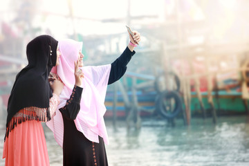 Obraz premium Two happy young muslim girls with Hijab making selfie photo on smartphone.Scene of morning with the mist and soft focus,fishing village