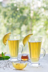 Still life of two tea cups with green tea, lemon, honey jar, peppermint and brew tea on white scratched background behind the window with summer green trees