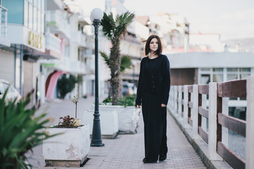 Fototapeta premium Young beautiful woman wearing total black clothes standing on the city embankment near sea