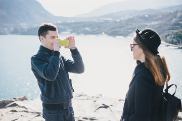 Man taking picture of his girlfriend using his smartphone near seaside and mountains. Travel together