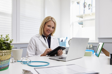 Female Doctor Sitting At Desk Using Digital Tablet In Office