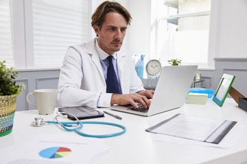 Male Doctor Sitting At Desk Working At Laptop In Office