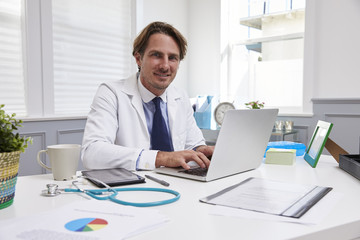 Fototapeta na wymiar Male Doctor Sitting At Desk Working At Laptop In Office