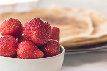 Fresh strawberries in white bowl with french pancakes creps in the background on Saturday morning breakfast, selective focus, healthy nutrition. Abstract family happiness concept 