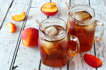 Two mason jar glasses of homemade peach iced tea on a rustic white wood background