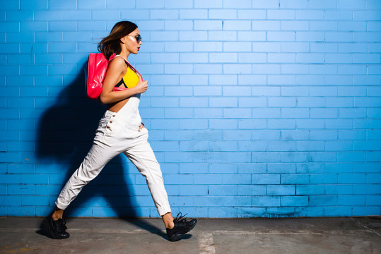 Beautiful young sexy hipster girl goes near urban blue wall background in yellow swimsuit, white jumpsuit, sneakers, sunglasses, pink backpack.