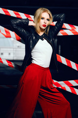 Fashion portrait of sexy blonde girl with red lips wearing a rock black style and red pants posing and smiling on the background of warning tape. Black, red, white.