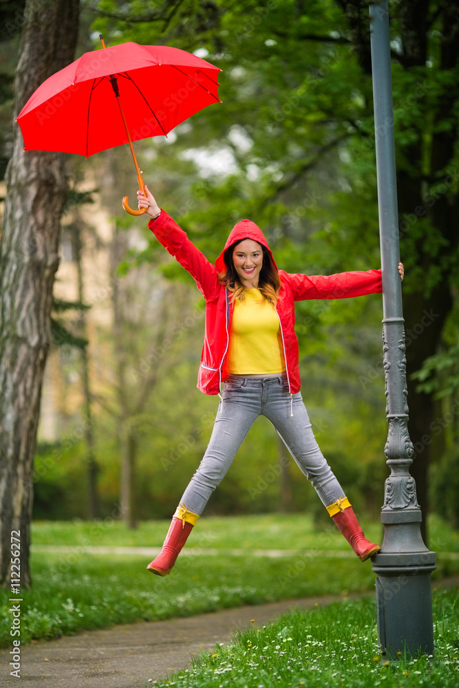 Wall mural Happy young woman is having fun on the rain. She climbed the outdoor lamp post in the park, smiling and looking at camera.