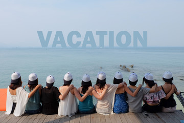 women friend group sit make arm hug hold around their friend's shoulder on wooden pier. They wear same design white and black color caps. looking at VACATION word on blue sea sky.
