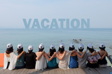 women friend group sit make arm hug hold around their friend's shoulder on wooden pier. They wear same design caps with FRIENDSHIP alphabets on each one. 
looking at VACATION word on blue sea sky.