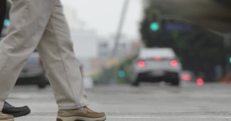 Focus on feet of pedestrians walking on edge of frame in streets in Downtown Los Angeles with passing traffic and other people soft focus in foreground. Recorded at 60fps with long telephoto lens
