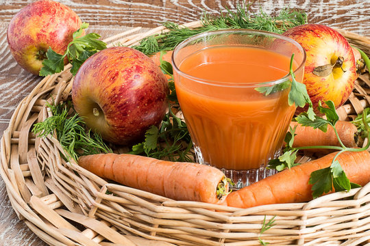   A glass with juice, apples and carrots.    A glass with juice, fresh apples and carrots in a wicker basket on a brown wooden background.