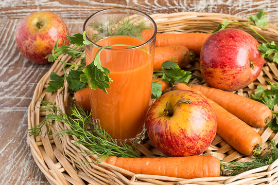   A glass with juice, apples and carrots .    A glass with juice, fresh apples and carrots in a wicker basket on a brown wooden background.