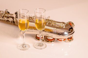 Shiny saxophone lying on white surface, musical notes paper and two glasses of champagne sitting next to it