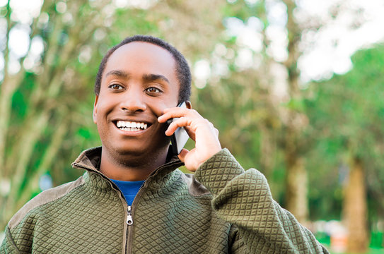 Handsome Hispanic Black Man Wearing Green Sweater In Outdoors Park Area Holding Up Phone To Ear And Talking Happily While Laughing