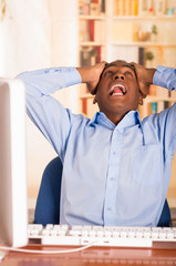 Young handsome man wearing blue office shirt sitting by computer leaning backwards in chair holding head in disbelief and frustration