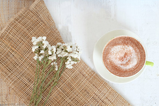 The glass of hot cocoa on white wooden and gunny sack background with white and yellow flowers
