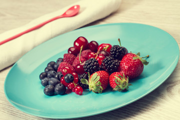 Frutos rojos, fresas, moras, frambuesas, arándanos, cerezas sobre un plato redondo celeste sobre una mesa. Vista de frente y superior