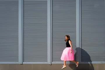 Beautiful sexy young hipster girl jumping outdoors.Posh woman with long brunette hair evening makeup wearing light fluffy pink rosy skirt and vans sneakers posing in the city urban style in summer