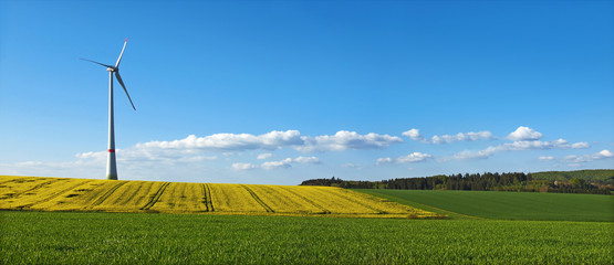 Landschaftliches Panoramabild mit Windrad, Rapsfeld und lockerer Bewölkung am blauen Himmel