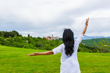 Back of woman with black hair raising hands and blurred ancient buildings and church on top of hill surrounded by trees on cloudy day, Labin, Croatia
