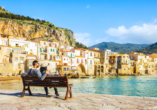 Couple sitting on bench, enjoying view of beach town of Cefalu in Sicily, Italy