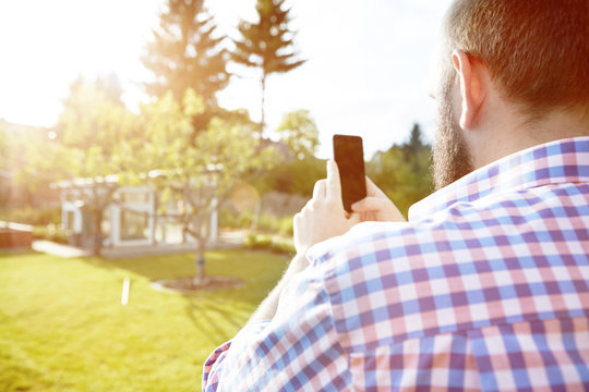 Young man with smartphone in garden, photografing his garden