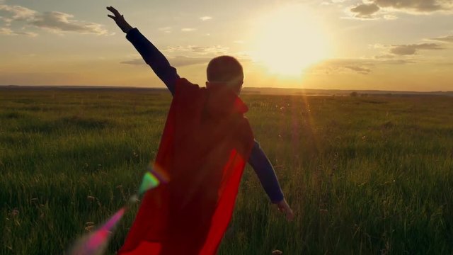 Boy dressed with a Superman cape running in a field, looking into the sunset