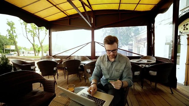 Handsome man in shirt and glasses using a laptop at cafe while holding credit card in the hands, on-line shopping outdoors. Copy space