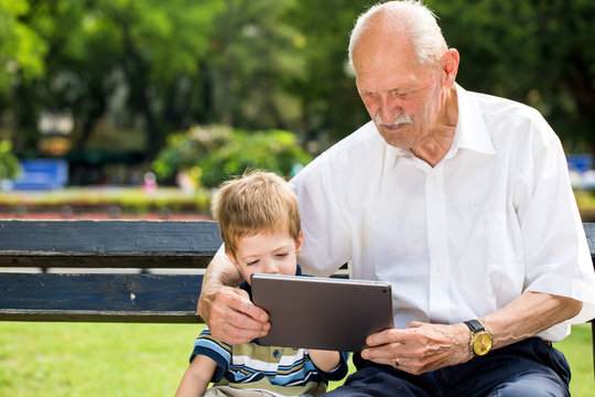 Grandchild teaching to his grandfather to use tablet on a bench in summer sunny day