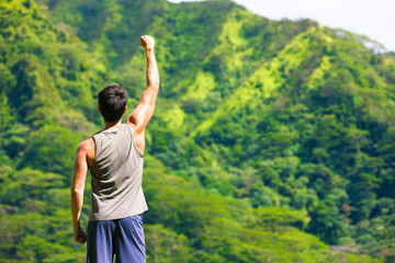 Strong fit male with fist in the air surrounded by lush green mountains.

