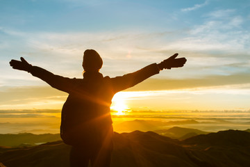Happy celebrating winning success woman at sunset or sunrise standing elated with arms raised up above her head in celebration of having reached mountain top summit goal during hiking travel trek.

