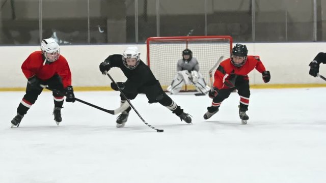 Four little hockey players skating towards the camera; boy in red uniform trying to get the puck from his opponent but falling down on ice