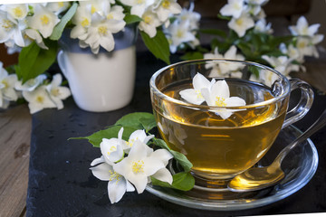 Transparent glass cup of tea with jasmine and blooming jasmine branch in blue ceramic cup on black board