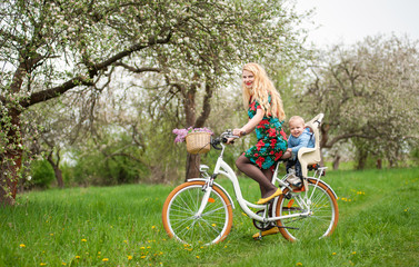 Beautiful female with long blonde hair in dress riding vintage bicycle with baby in bicycle chair, looking to the camera. In the basket lay a bouquet of lilacs, against spring garden