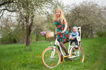 Fototapeta premium Mother with long blonde hair in dress riding bicycle with baby in bicycle chair. A bouquet of lilacs lay in the basket. Blooming fresh greenery in spring garden on the background