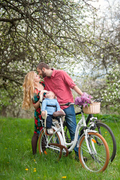Happy baby sitting in bicycle chair against loving parents on the background of blooming trees in spring garden