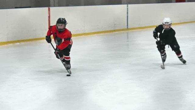 Two kids playing hockey on ice rink: one boy in red uniform handling puck and shooting it into the net