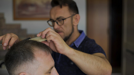 Obraz premium hairdresser cuts hair with scissors on crown client in professional hairdressing salon. The gray-haired man in the age of the client in the chair.