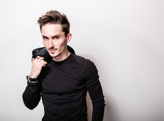 Attractive elegant man in black t-shirt and leather jacket in his hands posing on light gray studio background.